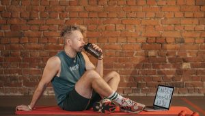 Athletic man sitting on exercise mat with water bottle, taking a break from workout, in a rustic indoor setting.